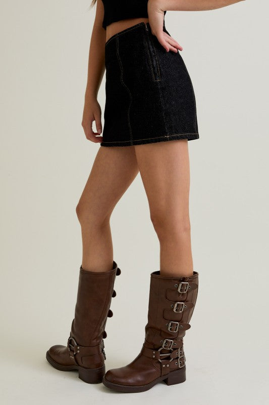 Person wearing a black denim skirt and brown knee-high boots on a plain background