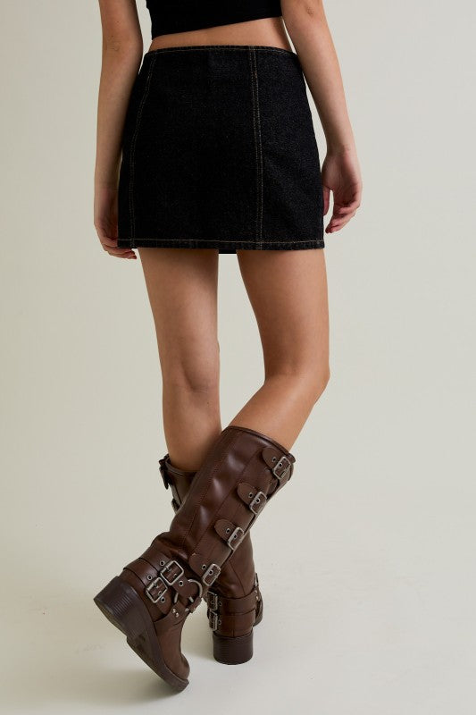 Person wearing a black denim skirt and brown knee-high boots on a plain background