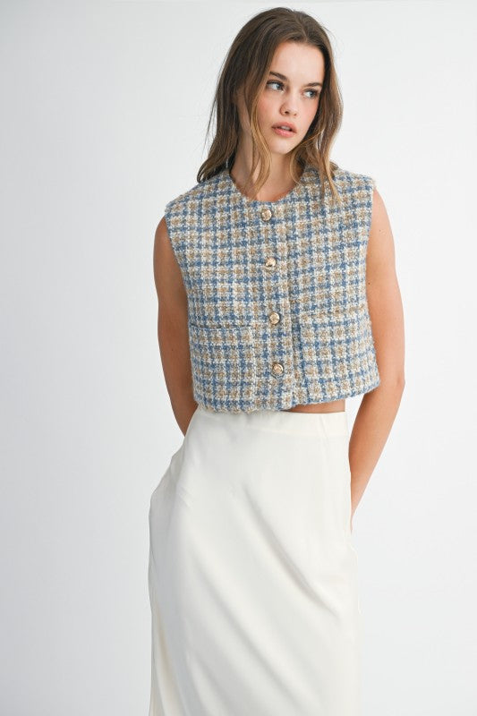 Woman wearing a patterned sleeveless top and white pants on a plain background