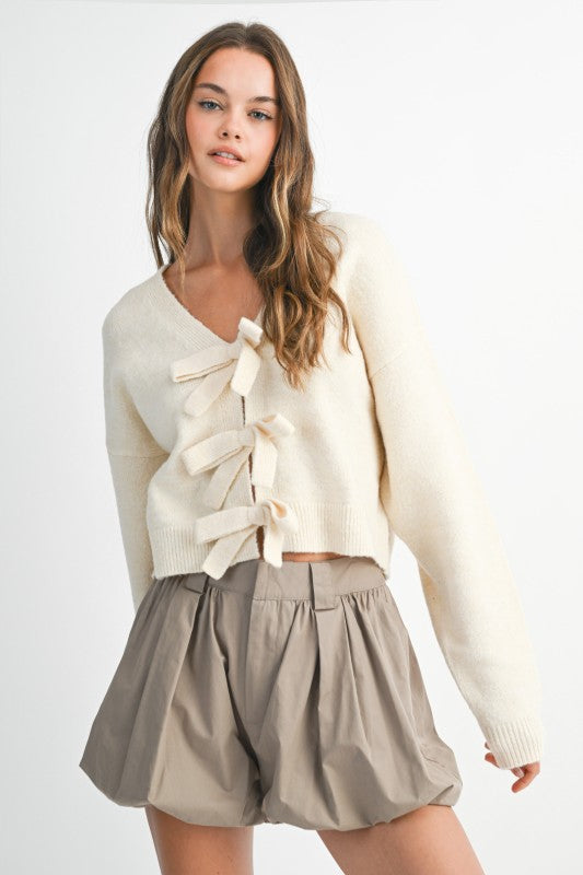 Woman wearing a cream sweater with a bow detail and beige shorts on a white background