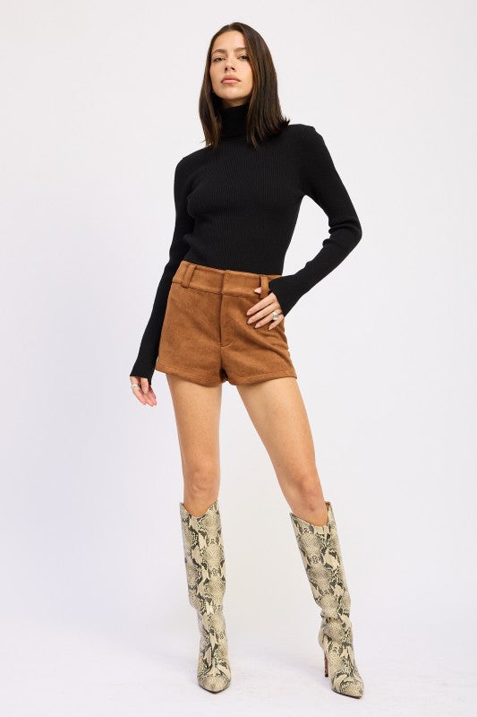 Woman wearing a black turtleneck, tan shorts, and patterned knee-high boots on a white background