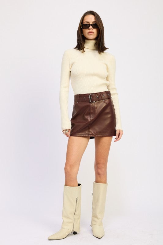 Woman wearing a cream turtleneck, brown leather skirt, and beige knee-high boots on a white background