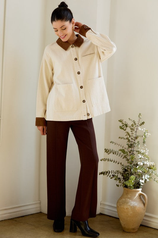 Woman wearing a cream jacket with brown collar and cuffs, standing indoors next to a vase with plants.