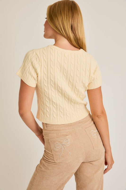 Woman wearing a beige cable knit top and beige pants with a bow detail on a plain background