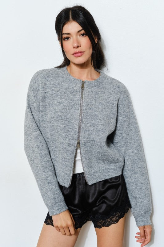 Woman wearing a gray zip-up sweater over a white shirt with black shorts on a light blue background