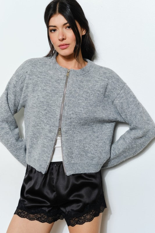 Woman wearing a gray zip-up sweater over a white shirt with black shorts on a light background