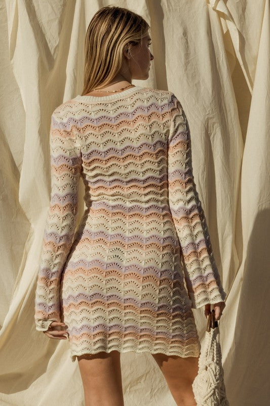 Woman wearing a colorful knitted dress standing against a white curtain.