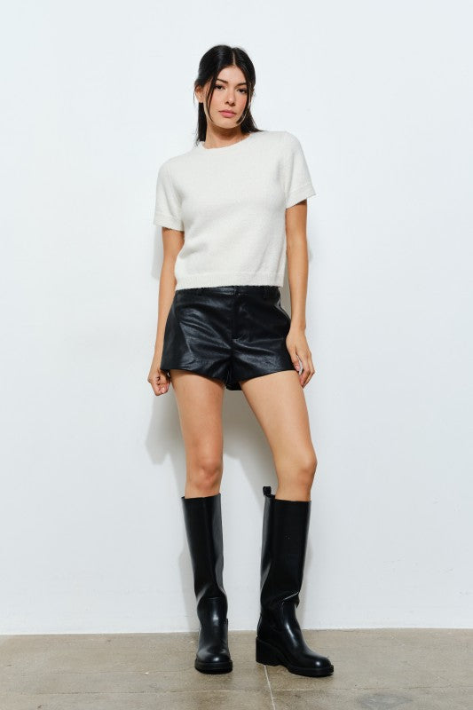 Woman wearing a white top and black leather shorts with black knee-high boots on a light background